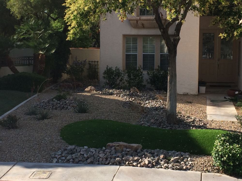 Low-maintenance front yard landscape with synthetic turf accent, decorative river rock dry creek bed, desert shrubs, and shade trees at a Las Vegas area residence by Southwest Greens of Nevada